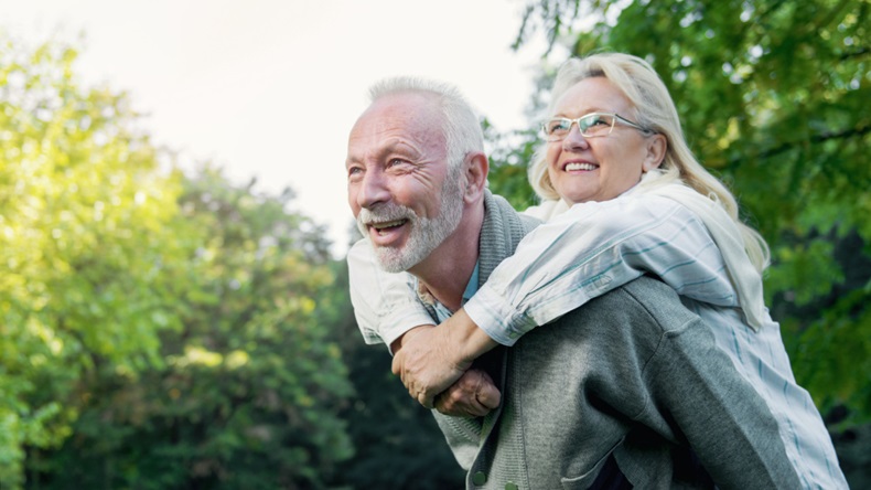 Happy senior couple smiling outdoors in nature, man giving woman a piggyback